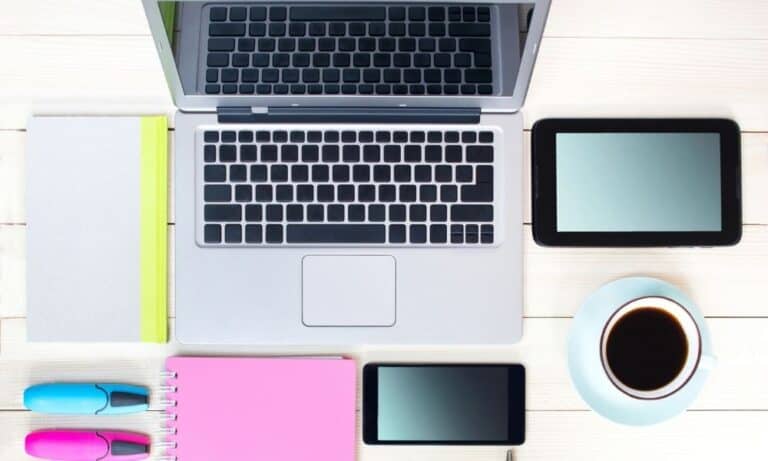 Overhead shot of desktop with open laptop, tablet, notebooks, highlighters, and a cup of coffee.