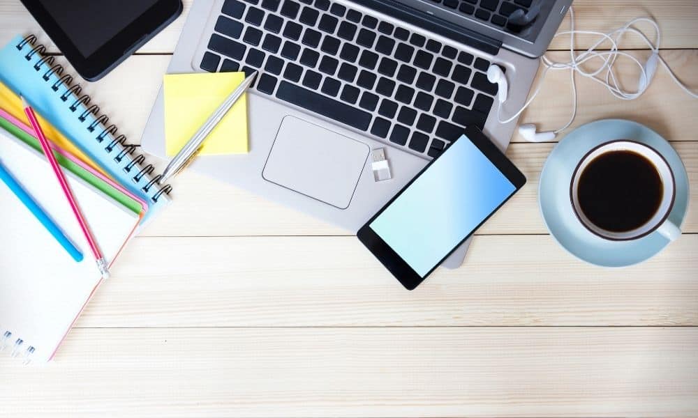 Overhead shot of desktop with open laptop, tablet, notebooks, pencils, and a cup of coffee.